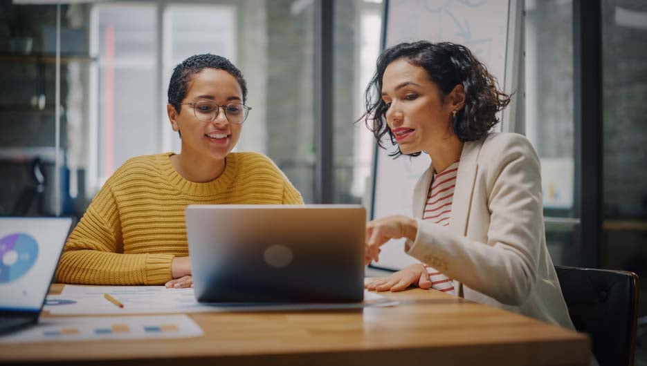 Two Diverse Multiethnic Female Have a Discussion in Meeting Room Behind Glass Walls in an Agency. Creative Director and Project Manager Compare Business Results on Laptop and App Designs in an Office.
